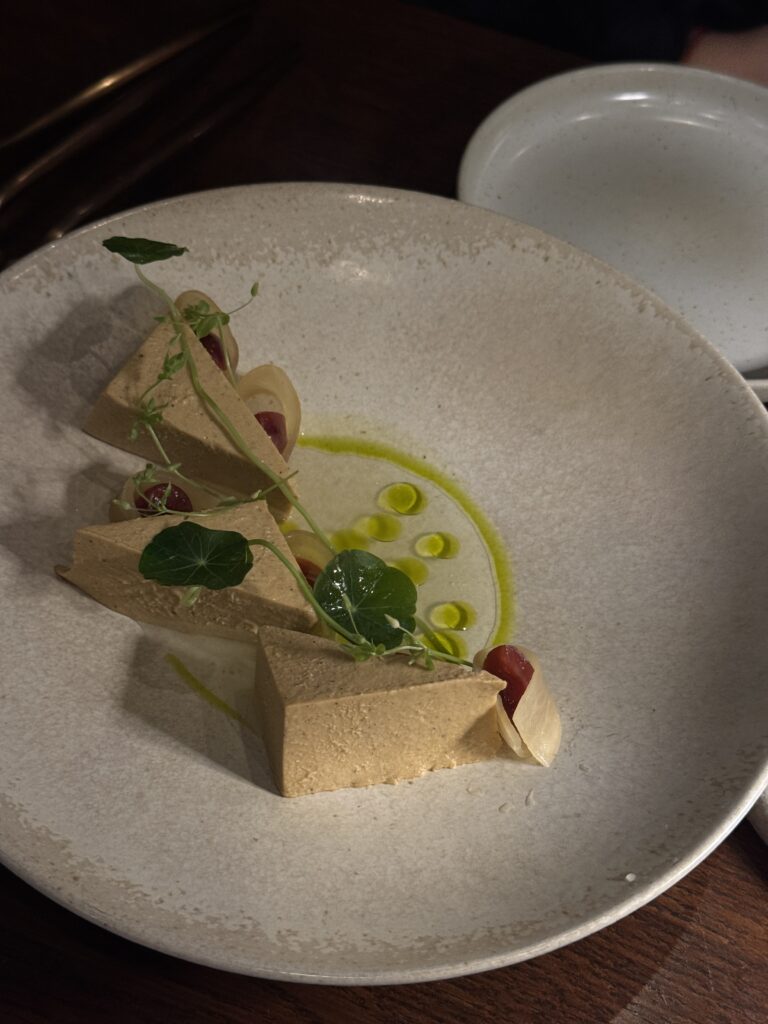 Fermentierter Tofu in dreieckigen Stücken, angerichtet mit Kräutern, Öltröpfchen und Gemüse auf einem hellen Keramikteller.